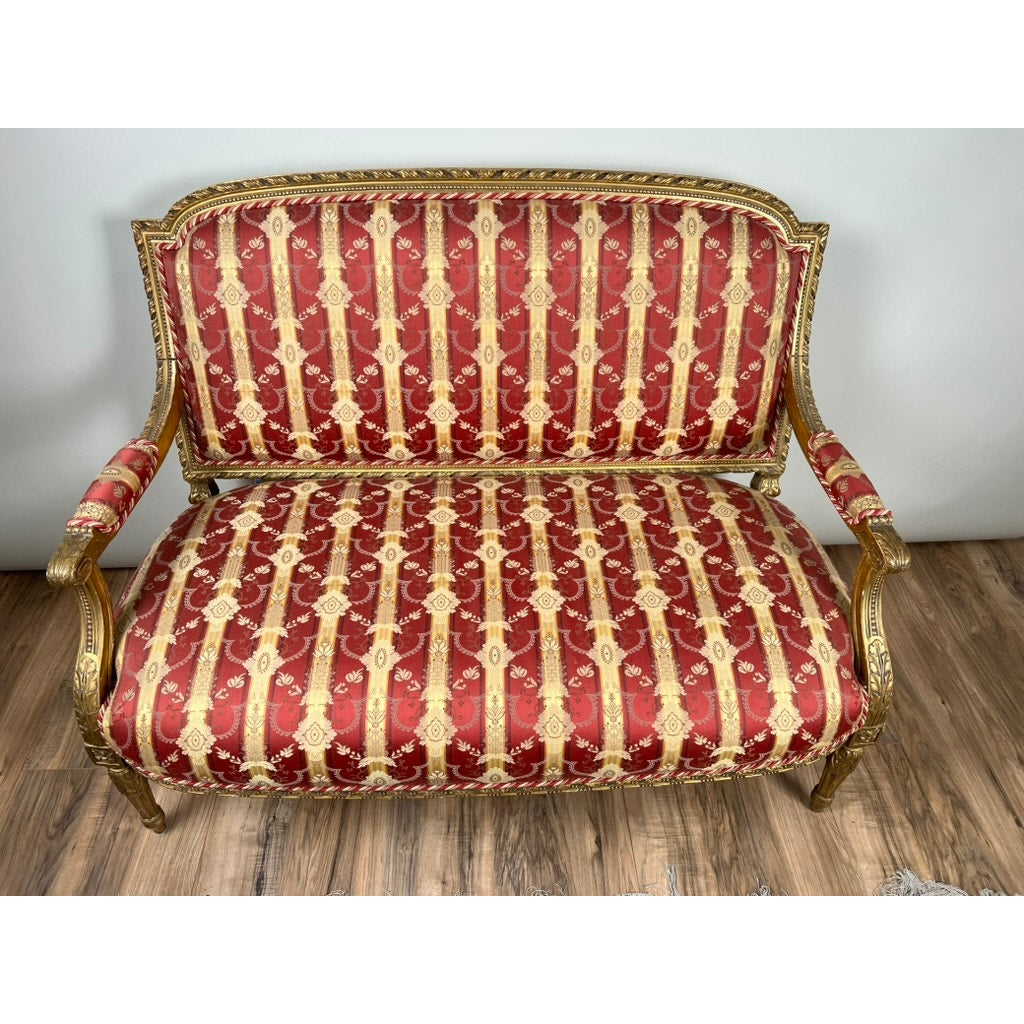 Antique Louis XVI style giltwood settee in silk damask c. late 19th century, featuring a gold ornate frame and red upholstery with gold floral and striped patterns, displayed on a wooden floor against a plain white wall.