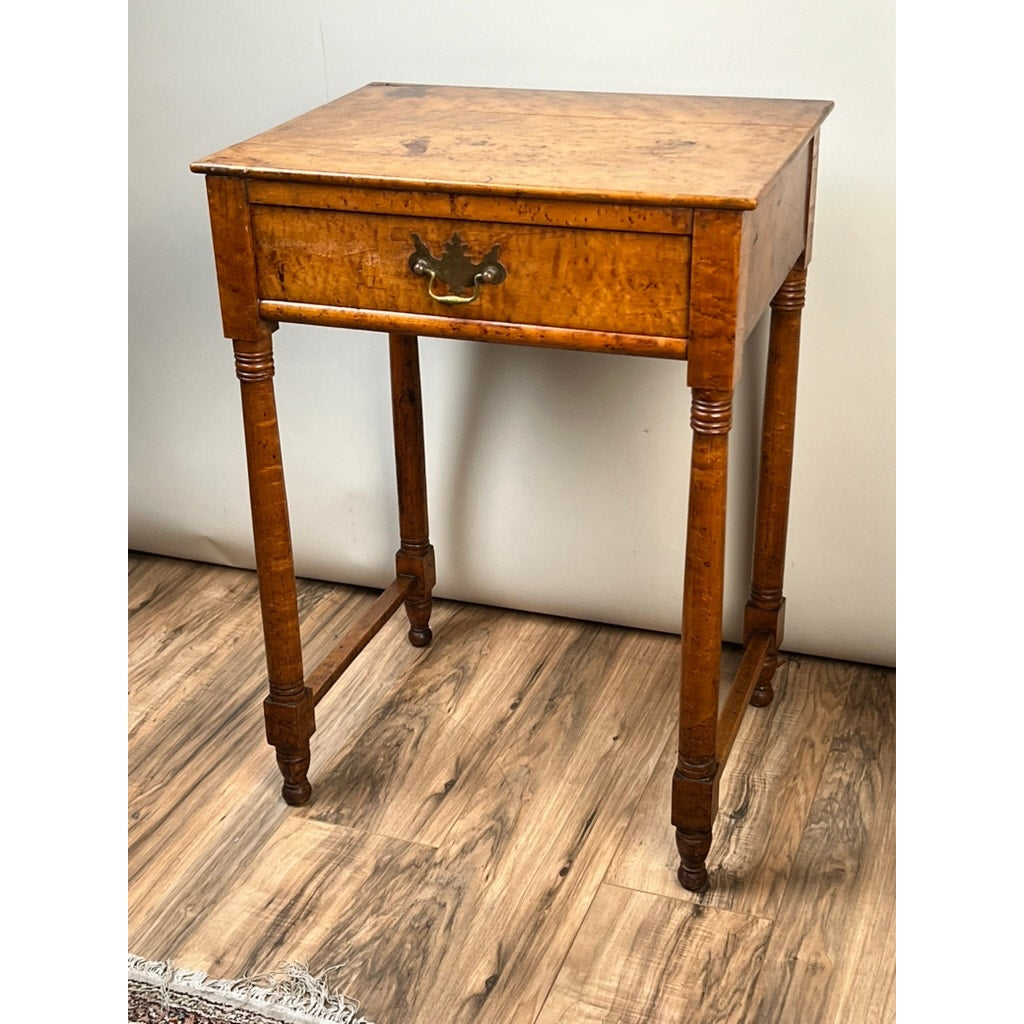 An Antique Sheraton Birdseye Maple One Drawer Stand, early 19th century, features a single drawer with brass handle and stands on four slender turned legs. Shown on a wood floor against a plain white wall. Brand: Unknown.