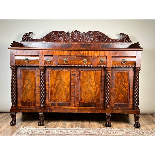 Elevate your space with this Antique American Empire Mahogany Sideboard, featuring rosewood edge banding and attributed to Philadelphia craftsmanship circa 1840. Offered by Pearl City Antiques, this elegant piece boasts ornate carved details, four drawers, four cabinet doors, claw feet, twisted columns, and a striking flame mahogany wood grain. Shown here set against a plain wall on a wooden floor with the edge of a rug visible.