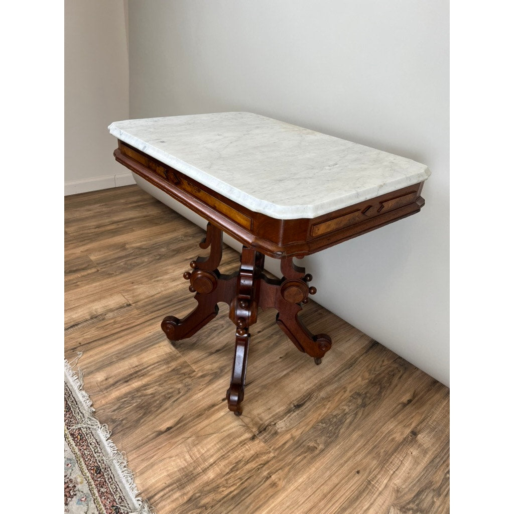 An Antique Eastlake Victorian Walnut Marble Top Stand, c. late 19th century, with an ornate base, stands against a plain wall on a wooden floor; part of a patterned rug appears in the bottom left. Brand: None/Unknown.
