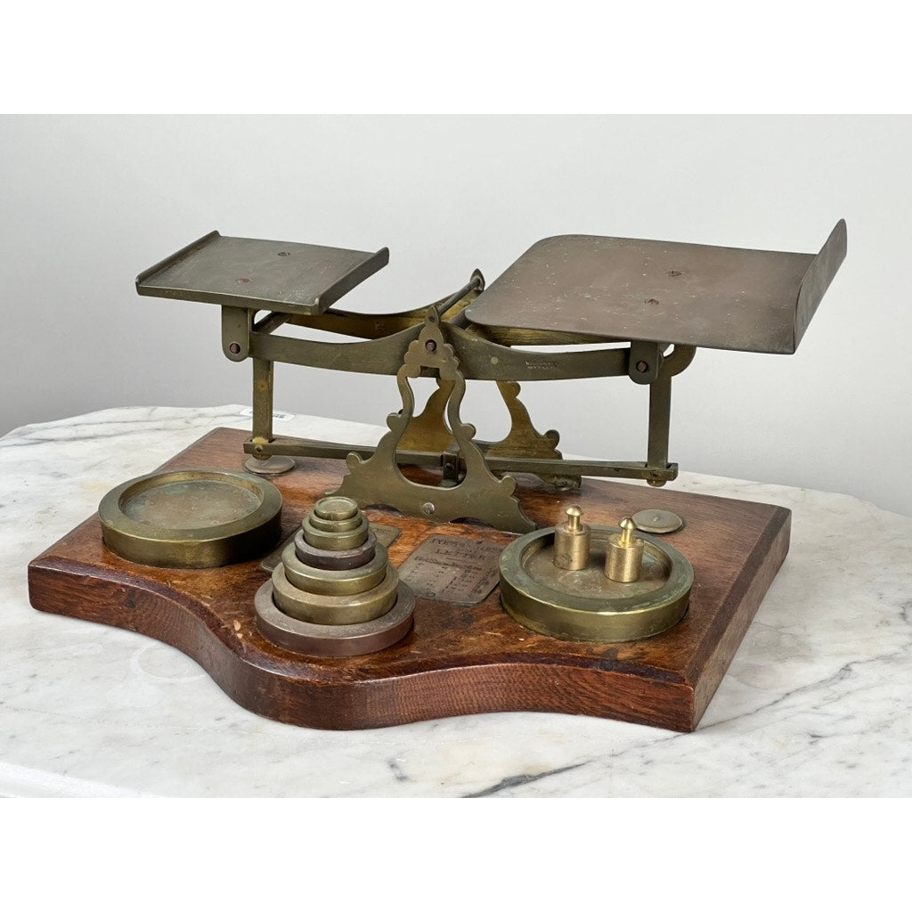 An Antique 19th Century English Postal Scale with two metal pans and brass weights rests on a wooden base atop a marble surface. Brand: Unknown.