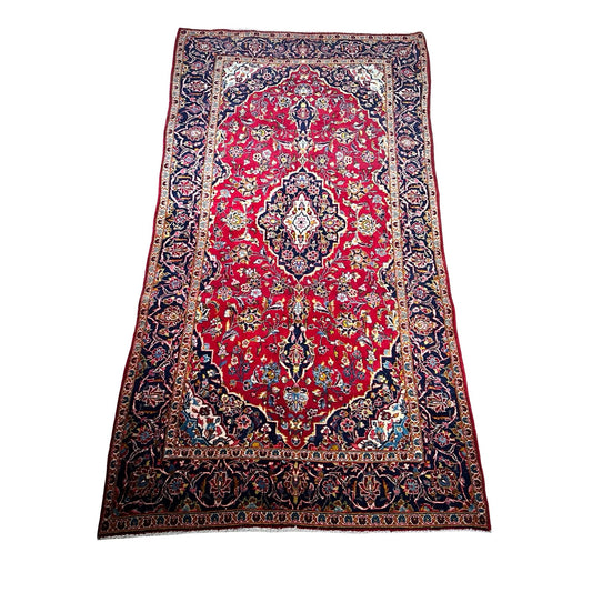 A vintage hand-knotted Persian Kashan wool gallery rug (c. 1950–1970, 4’5 x 8’8) features a rectangular design with a red field, intricate blue, ivory, and gold floral and geometric patterns, and a wide navy border with ornate details.