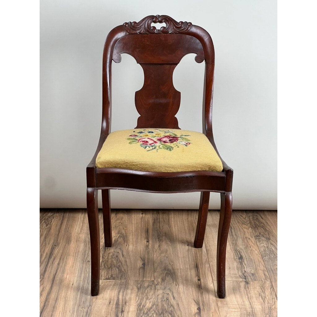An Antique American Empire Flame Mahogany Saber Leg Side Chair from the mid-19th century, with a carved backrest and yellow floral needlepoint upholstery, stands on a wooden floor against a plain light background.