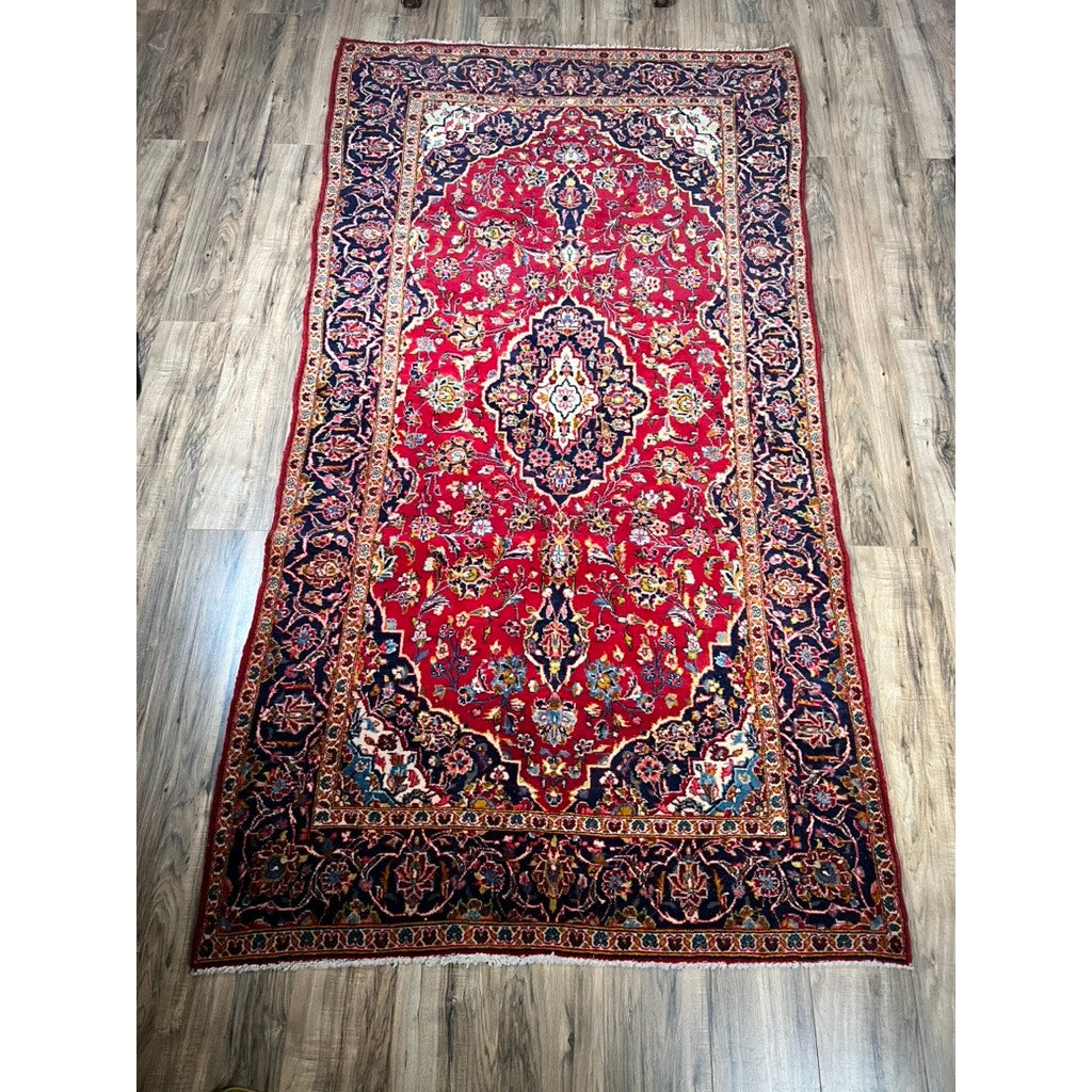 A vintage hand-knotted Persian Kashan wool gallery rug (c. 1950–1970, 4’5 x 8’8) with a red center and ornate patterns is displayed on a light wood floor. Brand: Unknown.