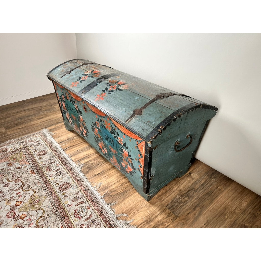 An Antique Swedish Oak Kista Dowry Chest c. 1841, featuring blue paint with orange and red floral accents and a curved lid, sits on a wooden floor by a patterned rug—a striking piece of Swedish folk art.