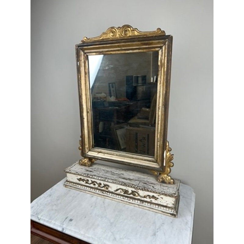 An Antique French Empire Gilt Wood Vanity or Table Mirror (c. Mid 19th Century) with an ornate gold frame rests on a marble-topped table, its elegant design standing out against the neutral wall backdrop.