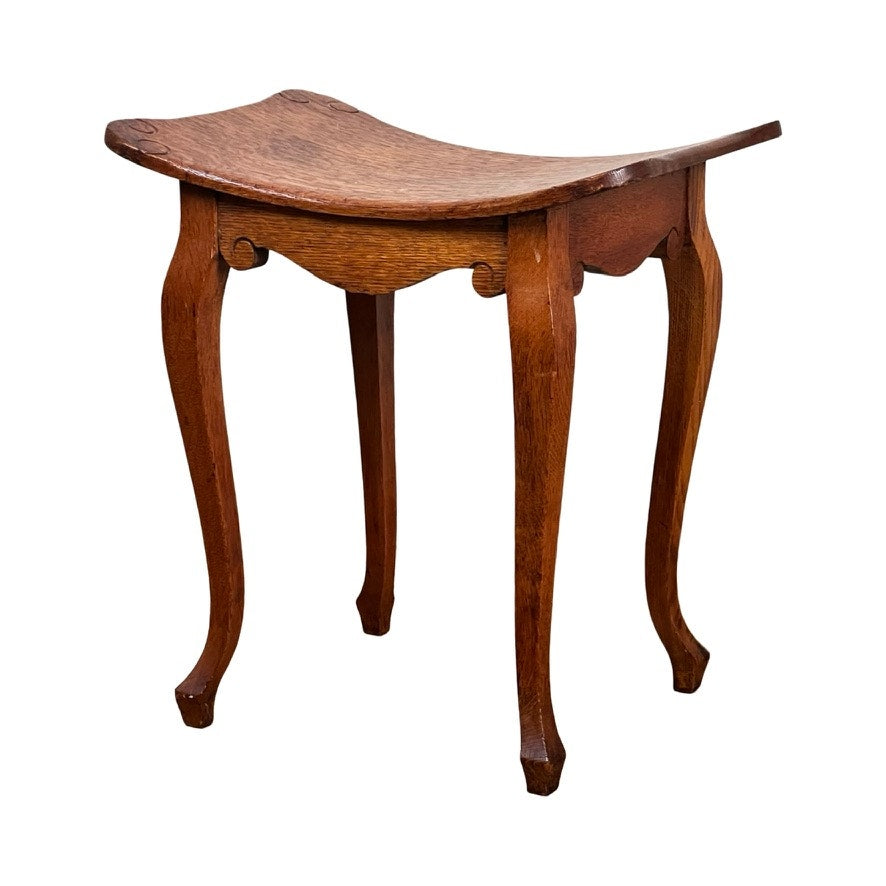 The Antique Arts and Crafts Saddle Shaped Footstool c. 1900 features a gently concave seat, curved legs, and a natural wood finish, offering an elegant traditional design reminiscent of vintage oak stools. Brand: None/Unknown.