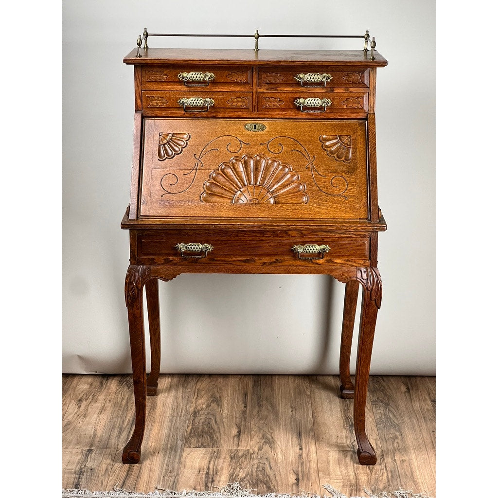 An Antique Victorian Oak Ladies Writing Desk c. 1900 stands on a wooden floor against a plain background, featuring ornate carvings, brass handles, a slanted front, four small upper drawers, and curved legs. Brand: Unknown.
