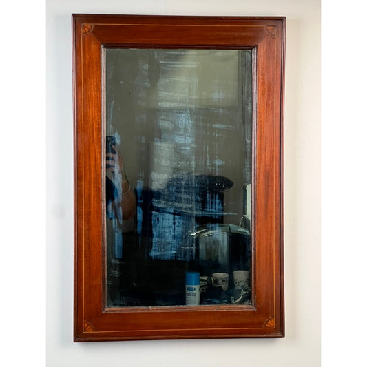 An American Federal Mahogany Wall Mirror with Fan Inlay, featuring its original hand-silvered glass (circa 1810–1825) from Pearl City Antiques, displays a timeworn wooden frame and softly clouded glass that reflects a hazy scene—a person’s hand holding a phone, various containers, and a white wall in the background.