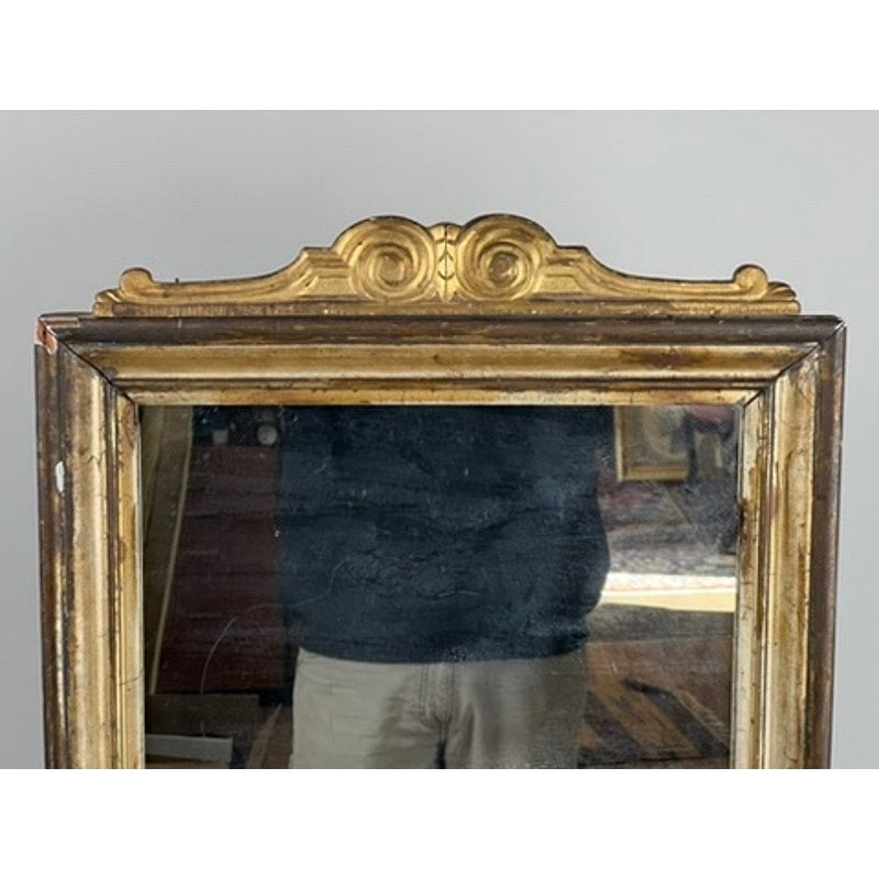 This Antique French Empire Gilt Wood Vanity or Table Mirror, c. Mid 19th Century, features an ornate gold crest on its wooden frame. The glass shows some wear, partially reflecting a person in a dark shirt and light pants.