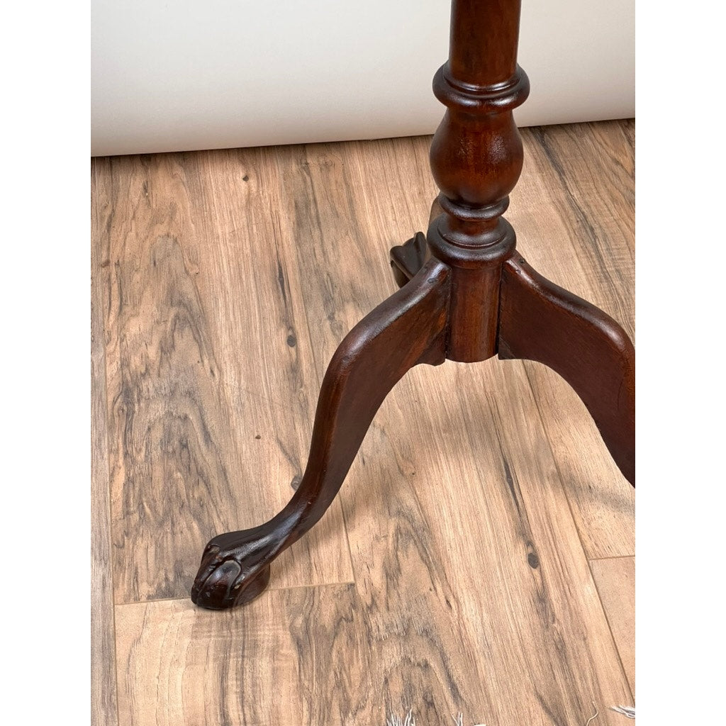 A close-up of a carved claw foot table leg from the Antique George III Tilt-top Tea Table (c. Early 19th Century) by Pearl City Antiques, crafted from dark polished wood with ornate curves and detailing, resting on a light brown wood floor.