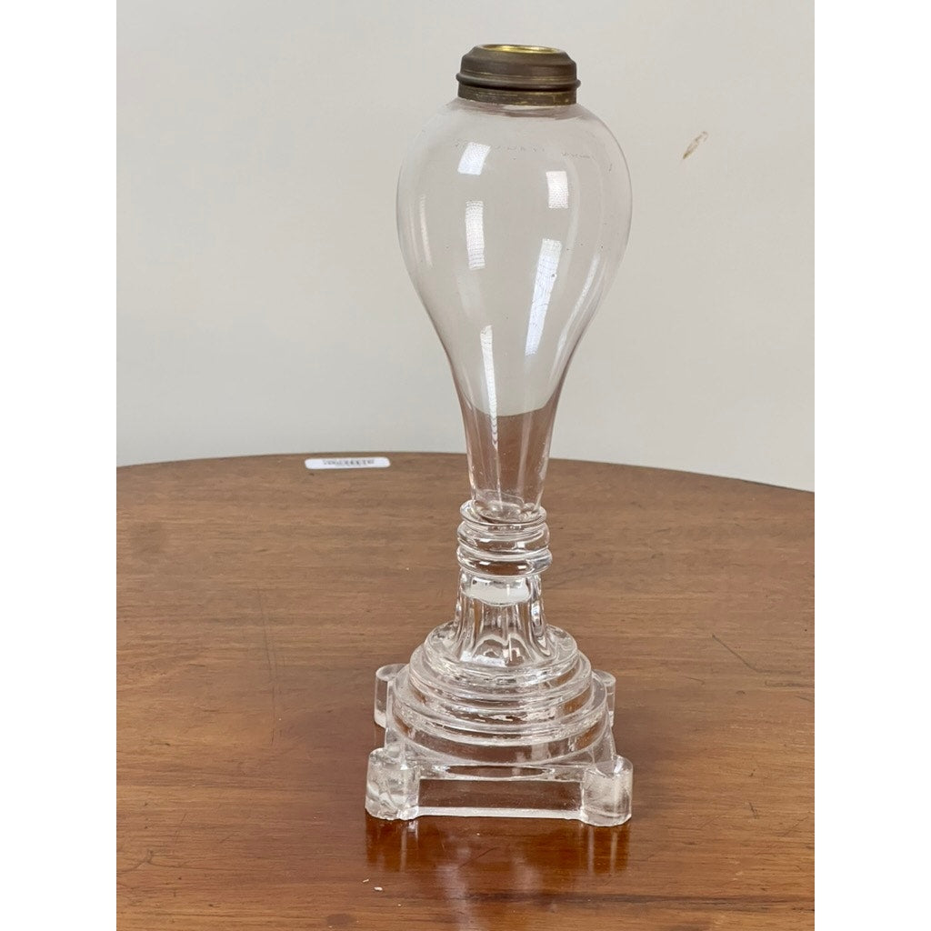 An Attributed Boston & Sandwich pressed glass boudoir fluid lamp, circa 1850, featuring a clear glass base with a round, bulb-shaped top and a square, stepped bottom, sits on a wooden table against a plain background—an elegant example of early American lighting.