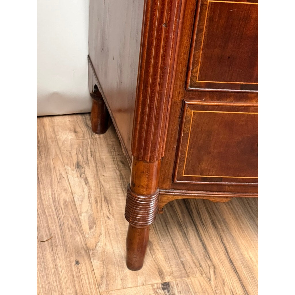 Close-up of the lower corner of an Antique Sheraton Mahogany Bowfront Chest (early 19th century) with decorative inlay, rounded legs, and ribbed details, shown on a light wood floor near a white wall.