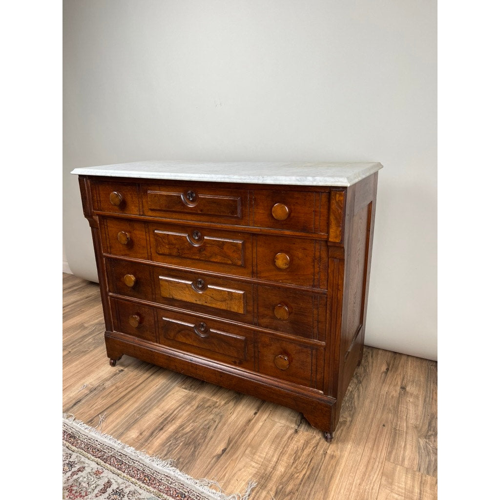 An Antique Victorian Marble Top Four Drawer Chest c. 1910 with round wooden knobs stands on a hardwood floor beside a patterned rug against a plain light-colored wall.