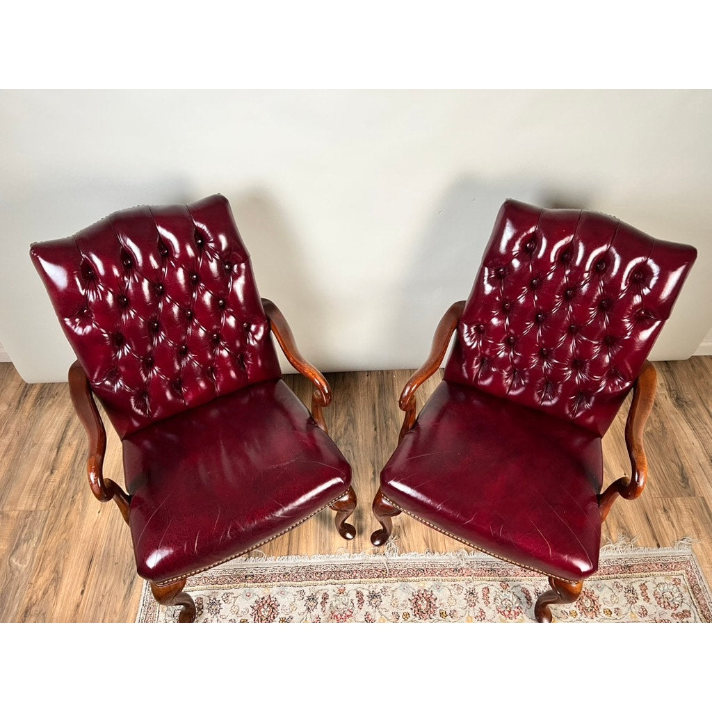 A pair of Chippendale Style Tufted Burgundy Leather Library Armchairs sit side by side on a wooden floor, partially positioned on a patterned rug against a plain, light-colored wall.