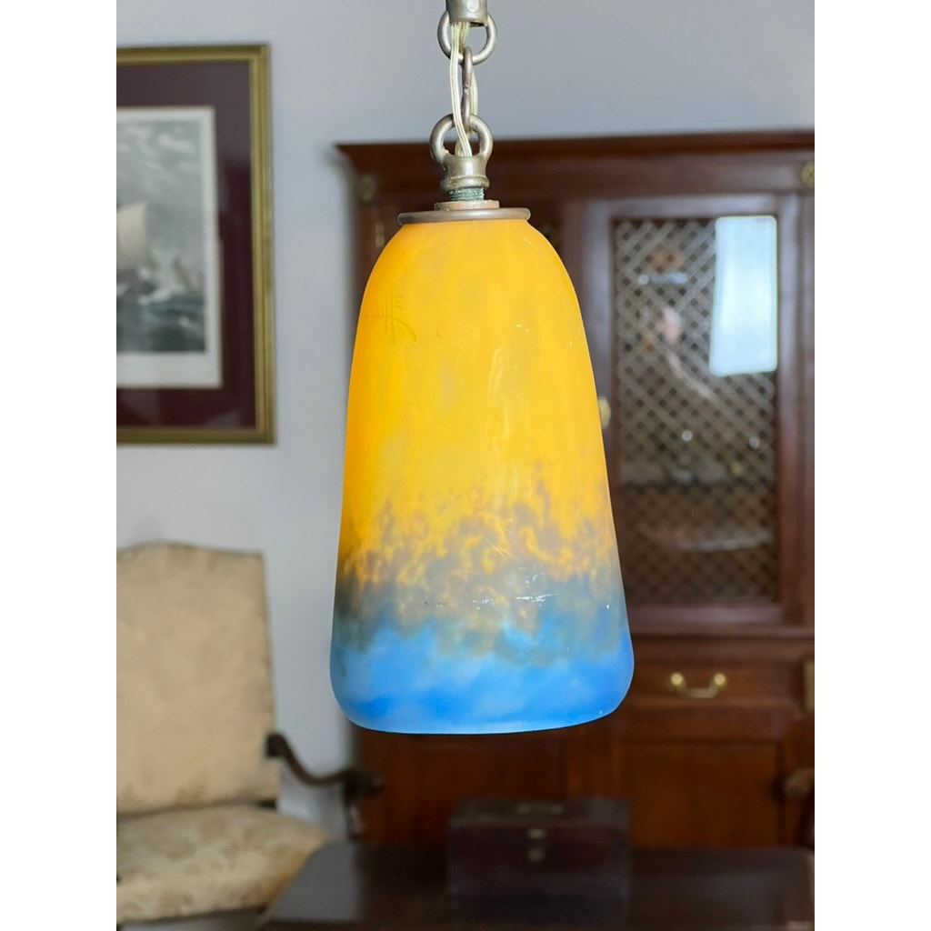 The Vintage Muller Frères Lunéville French Brass and Art Glass Four Light Chandelier, featuring a frosted Art Nouveau shade in yellow, gray, and blue tones, hangs by a chain before a wooden cabinet, framed art, and a beige upholstered chair.