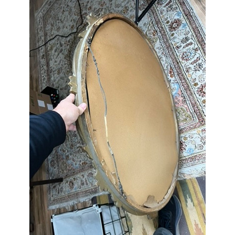 A person holds an Antique Gilt Wood Oval Mirror (early 20th century) with a damaged, cracked backing exposing wood. The frame rests on a patterned rug and wooden floor amid assorted objects. Brand: None/Unknown.