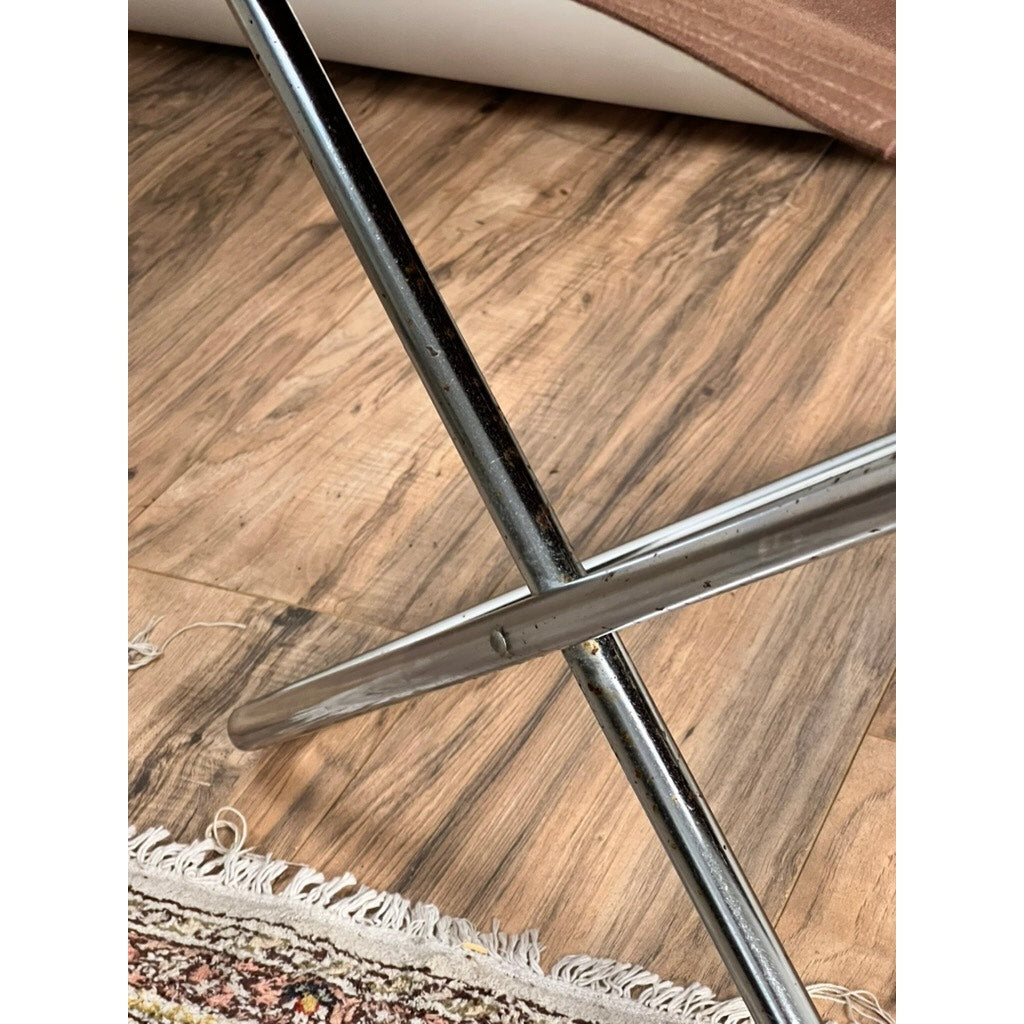 Two intersecting chrome rods arranged in an X shape on a wooden floor, accompanied by the corner of a patterned rug and a rolled-up mat in the background, capture the minimalist elegance of Japanese modern design characteristic of the Vintage Uchida Z Chair Japanese Modern Chrome Sling Lounge Chair with Headrest by Uchida.