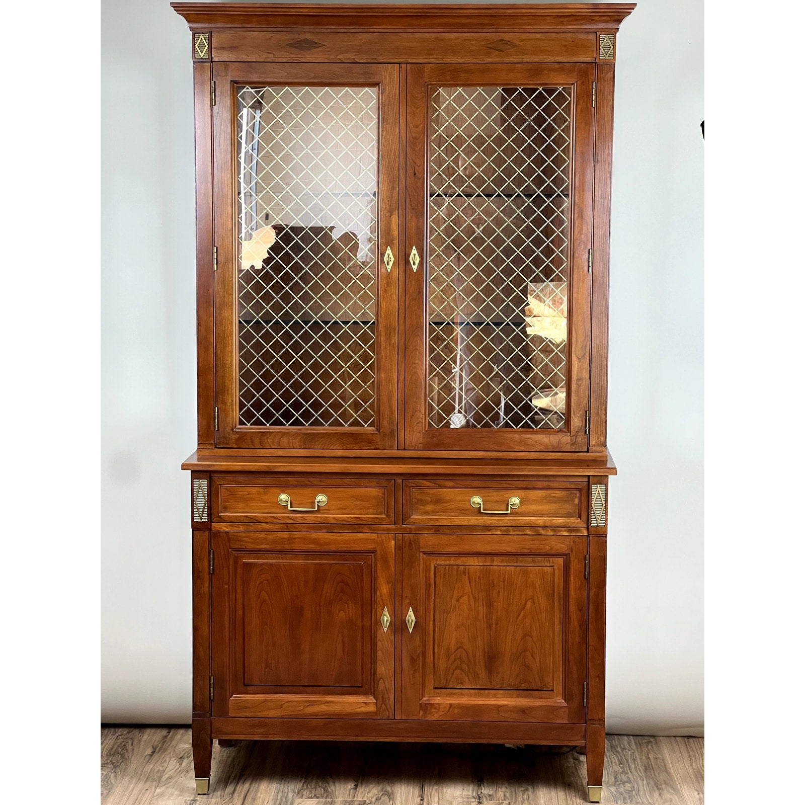 The Vintage William IV Style Cherry Glass Door Bookcase by Stickley features upper glass doors, lower drawers, solid wood construction, and brass accents, making it a classic addition to any room.