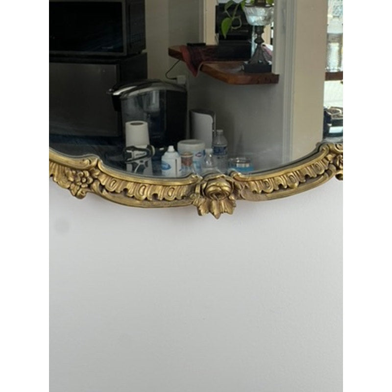 An Antique Gilt Wood Oval Mirror (early 20th century) with floral detailing reflects a kitchen counter holding a coffee maker, cups, water bottles, and paper towel rolls. Brand: Unknown.