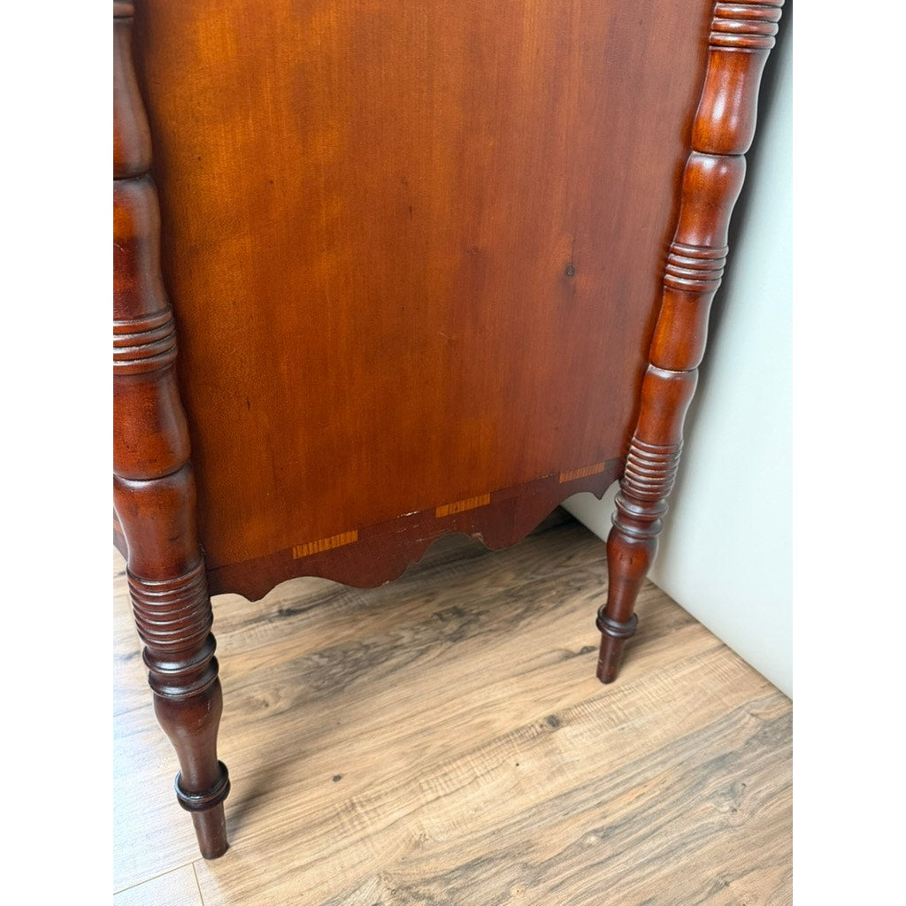 Close-up of an Early 19th Century New England Federal mahogany bow-front chest of drawers with backsplash, c. 1820–1840, featuring turned legs and a decorative bottom edge, standing on a light-colored wood floor next to a white wall.