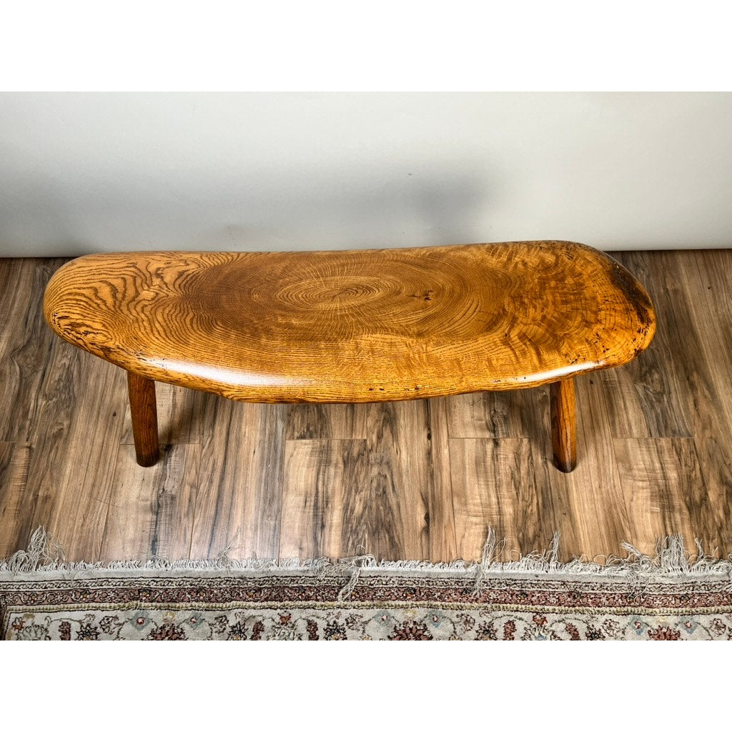 The Vintage Hand Made Oak Wabi Sabi Bench signed Neil Panyik features a smooth, oval seat and three round legs. Its natural polished finish highlights the oak grain, and it sits on a wood floor next to a patterned rug. Brand is unknown.