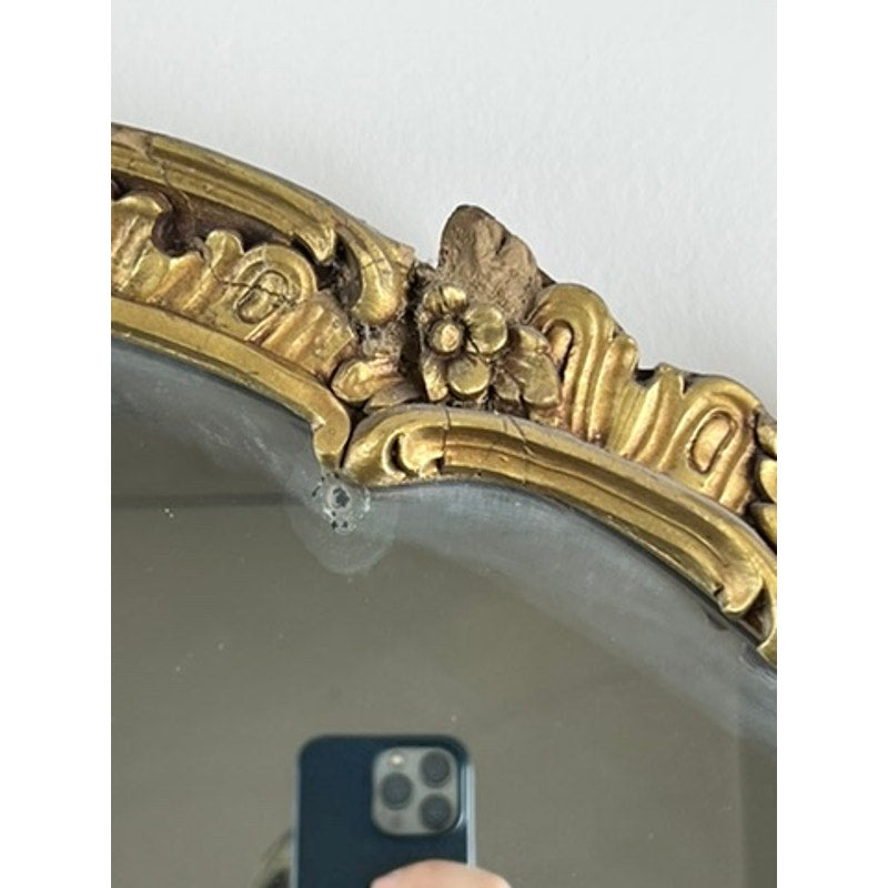 A person takes a photo of an Antique Gilt Wood Oval Mirror (c. Early 20th Century) with an ornate gold frame, floral top detail, and a small chip on the edge. Brand: Unknown.