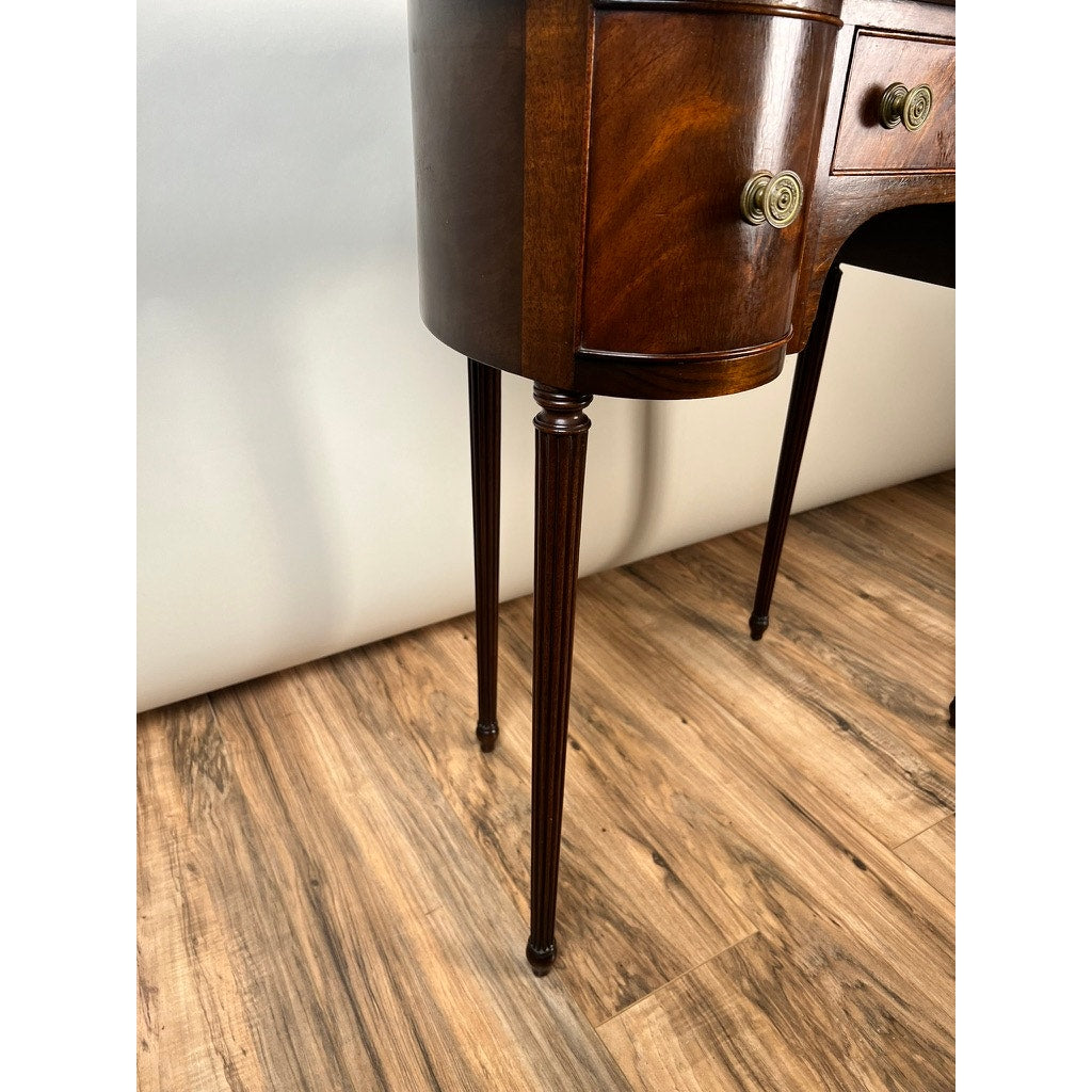 Close-up of a Vintage Edwardian Sheraton Revival kidney-form dressing table c. 1940, featuring mahogany veneers, curved drawers, brass handles, and slender tapered legs on a wood-patterned floor against a plain white wall.