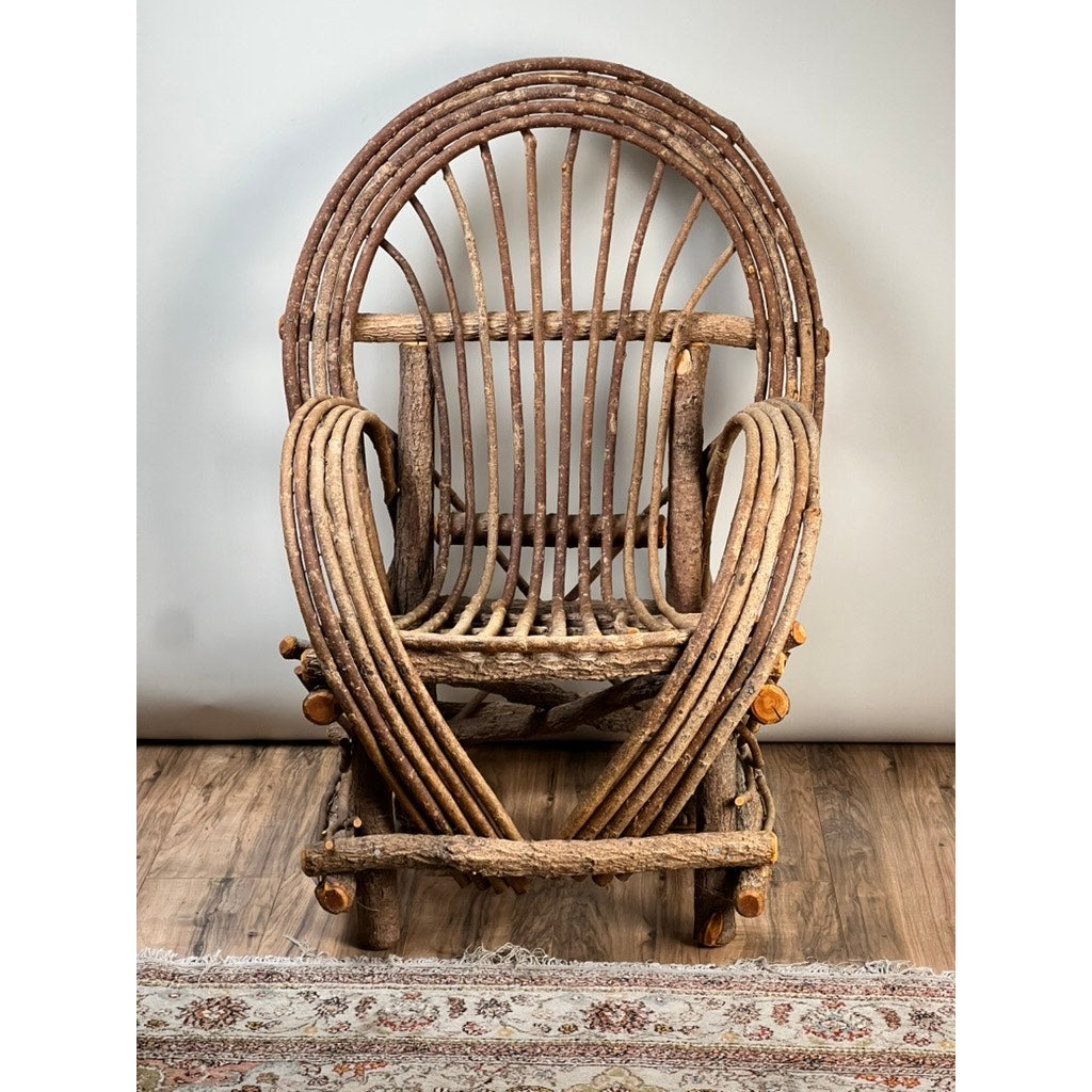 The Vintage Adirondack Bent Willow Arm Chair, crafted from curved unfinished branches, features a round back and looped armrests, bringing rustic cabin charm to any room. It sits on a wooden floor with a patterned rug below.