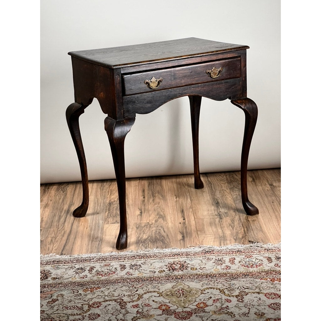 An Antique Queen Anne Style George III Oak One Drawer Stand (late 18th century) stands on a wooden floor beside a patterned rug, placed against a plain light-colored wall.