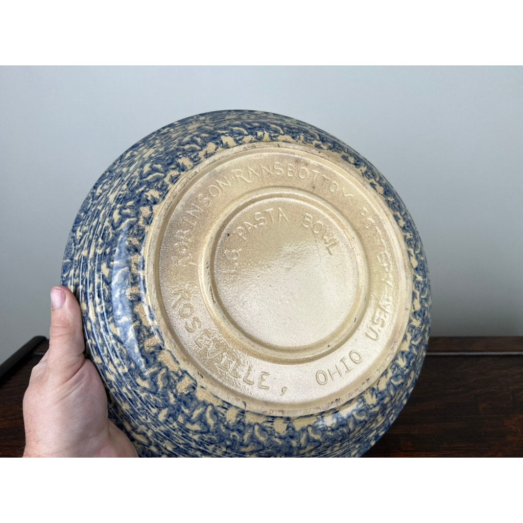 A hand holds an upside-down Robinson Ransbottom Roseville Blue Spongeware Pasta Bowl, revealing embossed branding and size details on its base—an iconic piece of vintage American ceramic craftsmanship.