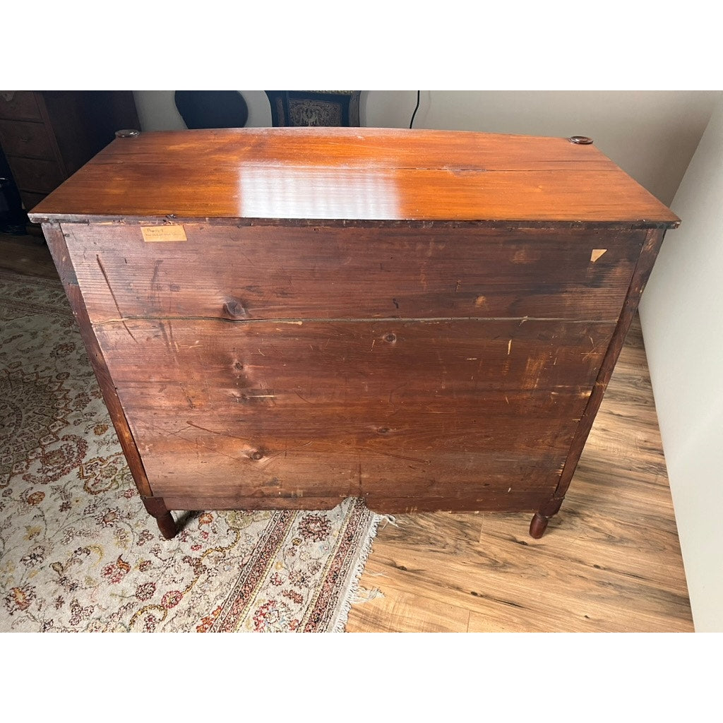 An Antique Sheraton Mahogany Bowfront Chest from the early 19th century stands on a wooden floor beside a patterned area rug and a cream-colored wall, its polished top contrasting with the more rustic, unfinished back.