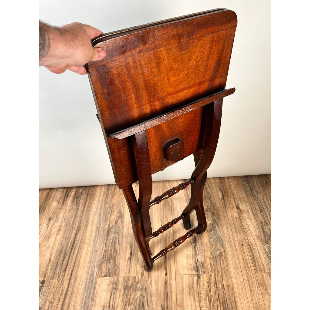 A hand holds the top of an Antique English Regency Mahogany Folding Coaching or Sporting Table (c. 1825–1845), partially open on a wood floor against a plain white background, showcasing its decorative leg detailing.