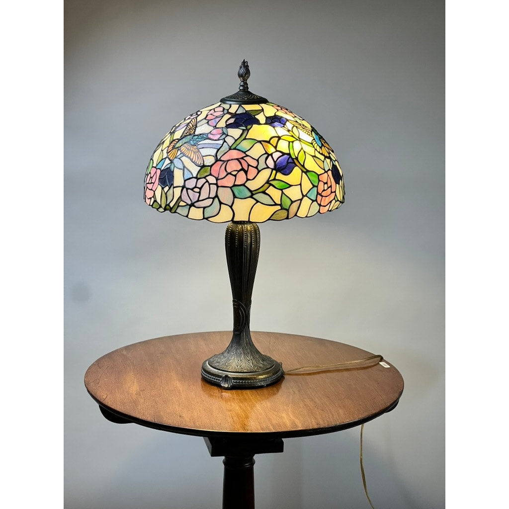 The Vintage Tiffany Style Floral Stained Glass Table Lamp sits illuminated on a round wooden table against a light gray background, casting a warm glow. Brand: None/Unknown.