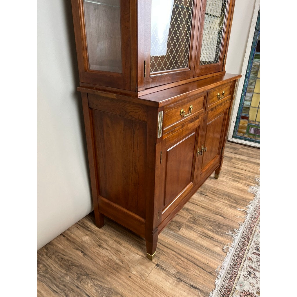 A Vintage William IV Style Cherry Glass Door Bookcase by Stickley with glass-paneled doors and metal accents stands on a wood floor near a patterned rug and a stained glass window.