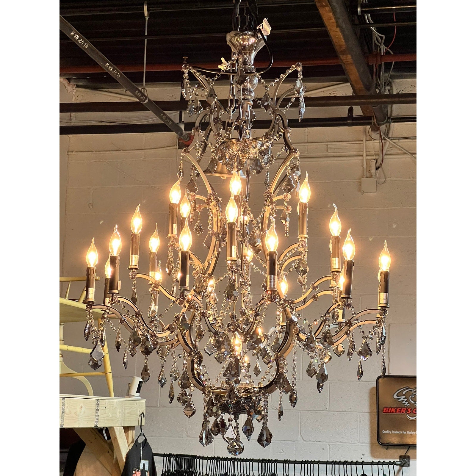 The Large Vintage Smoke Crystal 24 Light Chandelier hangs from the ceiling in an industrial space with exposed pipes and a white brick wall, its tiers of illuminated crystals creating a striking focal point.