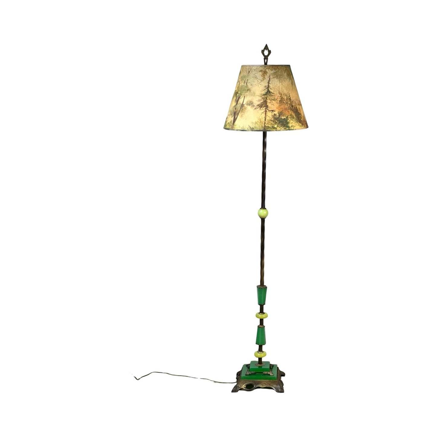 The Antique Art Deco Uranium and Slag Glass Lamp c. 1920 features a painted green and brown base, slender pole, and a beige lampshade with a tree scene. It is shown plugged in with a visible cord on a white background. Brand: Unknown.