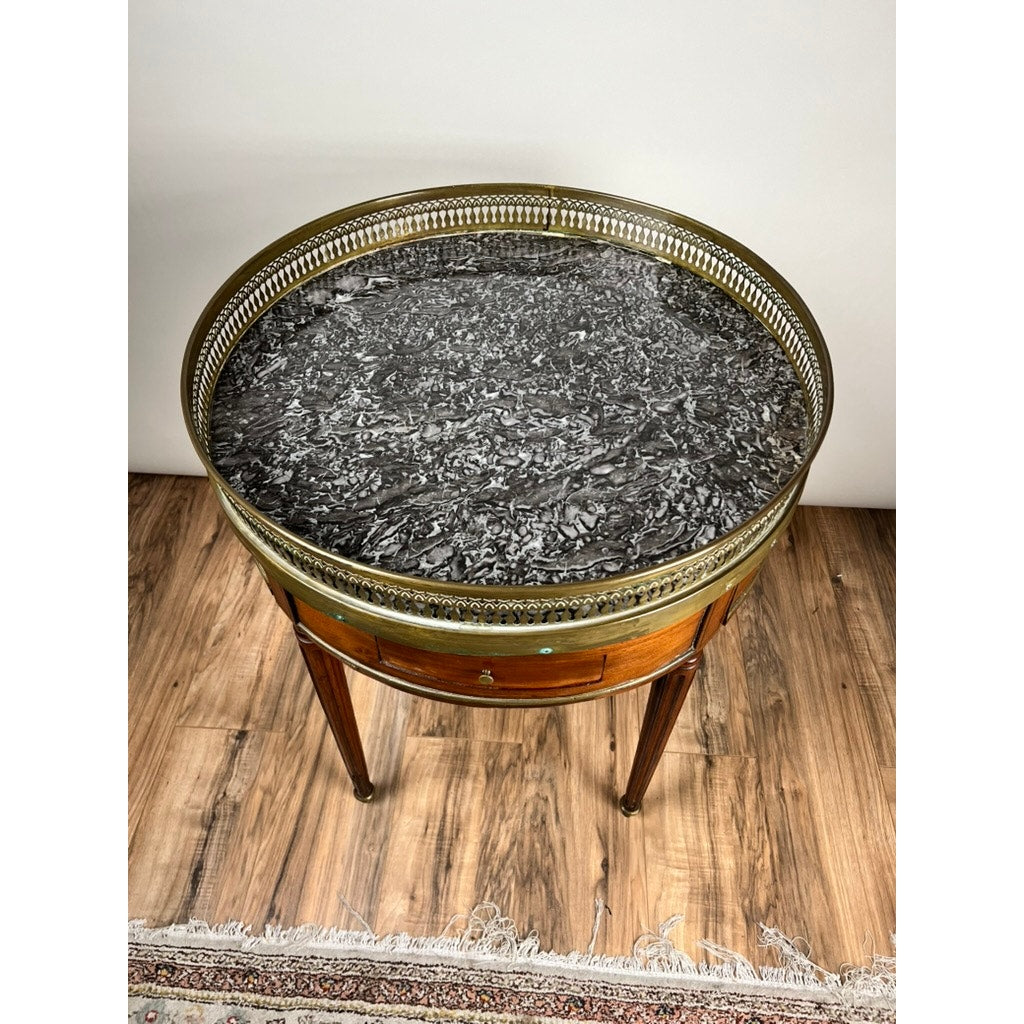 An antique 19th-century French Louis XVI-style mahogany bouillotte table with a marble top and pierced brass gallery stands on a wood floor beside a fringed rug.