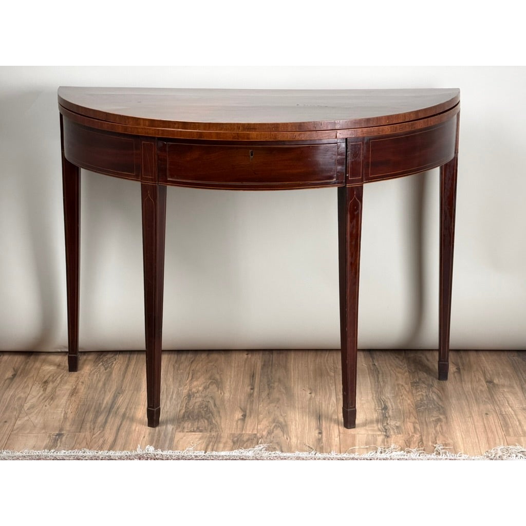 An English George III Mahogany and Satinwood-Inlaid Demilune Games Table, featuring a semi-circular shape with a single drawer and tapered legs.