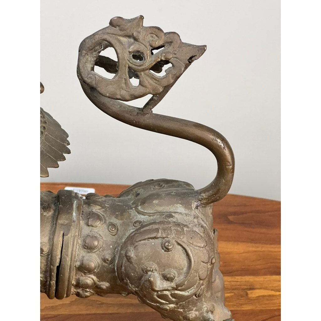 A close-up of a Vintage 17” Cast Brass Pixiu Tibetan Lion (c. 1960), featuring a stylized animal with a long curved neck and intricate dragon-like head, set on a wooden surface against a plain background. Brand unknown.