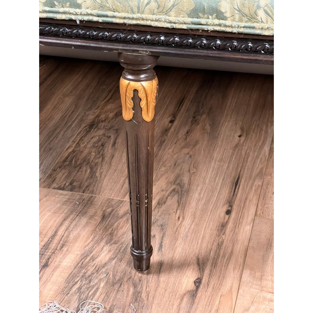 A close-up of an ornate wooden leg with gold accents, set on a wood floor beneath a Vintage Louis XVI Style Settee in green damask upholstery.