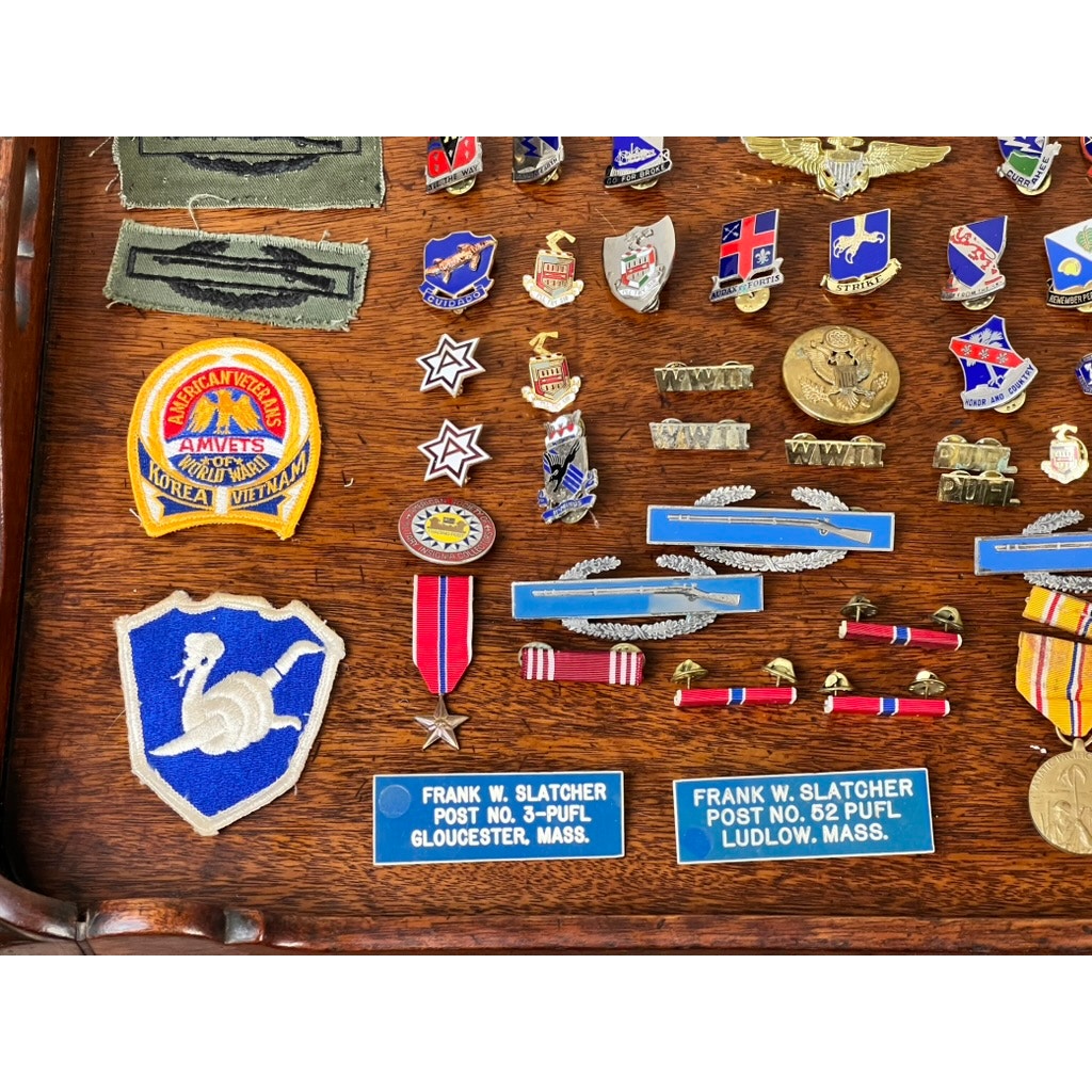 An Attributed WWII 158th Infantry Bushmasters Grouping of Frank W. Slatcher, Company I, Pacific Theater, features memorabilia such as patches, medals, pins, and nameplates in a display case on a wooden background.