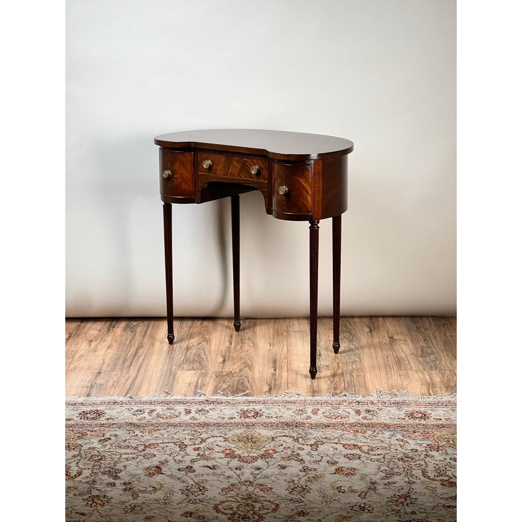 A Vintage Edwardian Sheraton Revival kidney-form dressing table c. 1940, with mahogany veneers, four slender legs, brass drawer pulls, and a curved front, stands on a wood floor with a patterned rug against a plain light background.