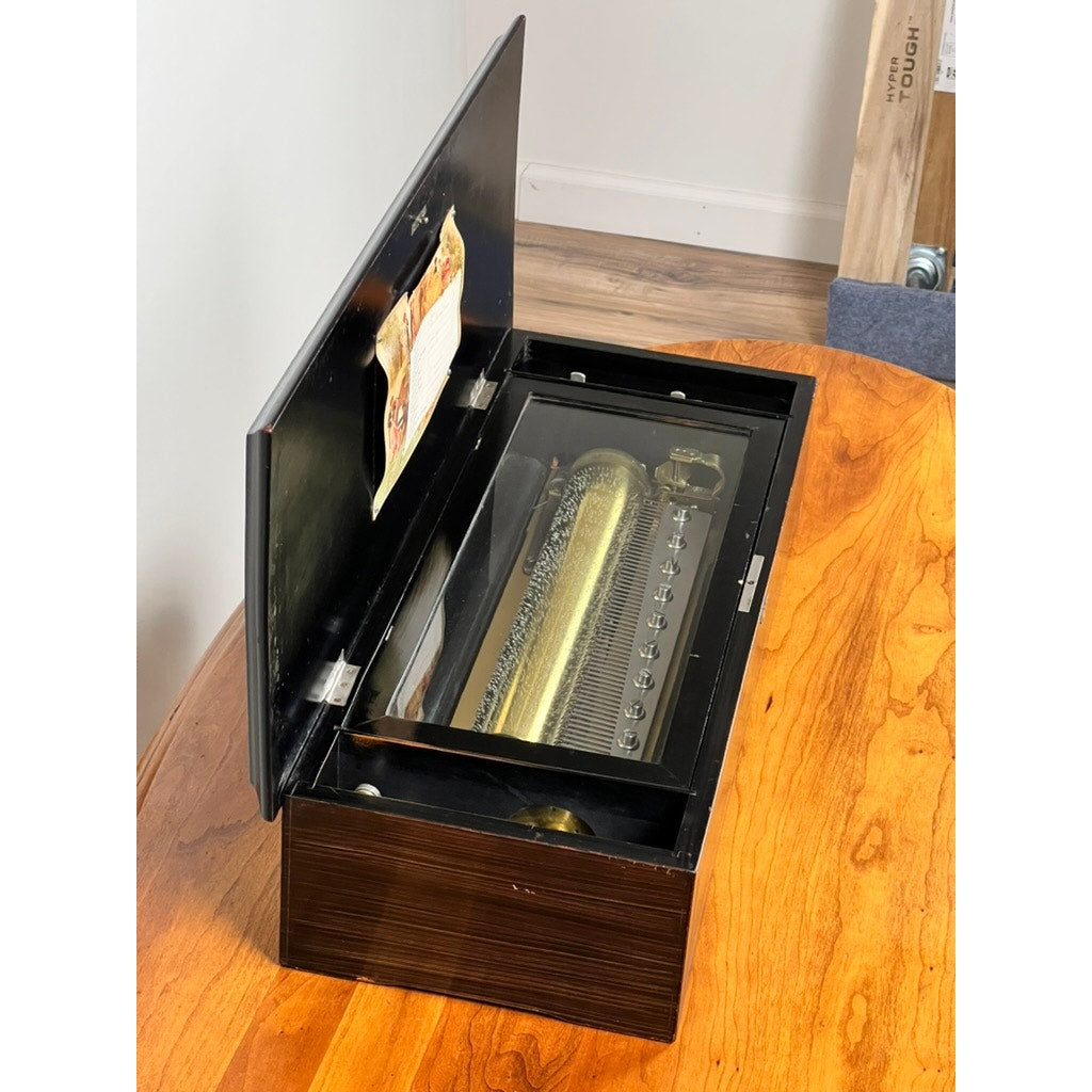 An Antique Swiss 12 Tune Music Box from the late 19th century, with visible combs and a cylinder mechanism, is displayed open on a polished wooden surface near a white wall, reflecting Victorian-era charm. Brand unknown.