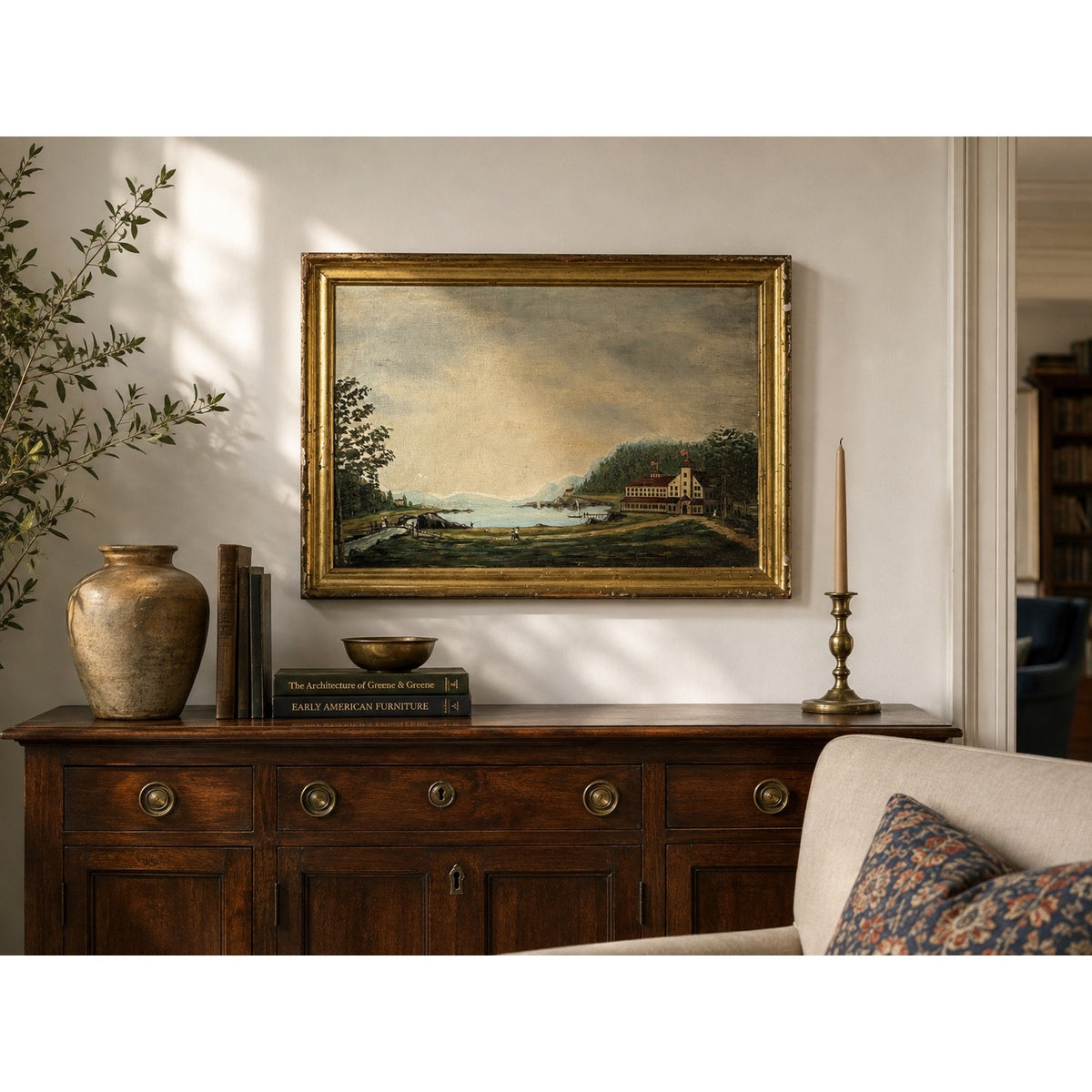 A wooden sideboard adorned with books, a vase, bowl, and candlestick sits beneath the framed 19th Century American Folk Art Adirondack or New England Resort Hotel Oil Painting from Pearl City Antiques on a white wall, next to a beige sofa accented with a patterned pillow.