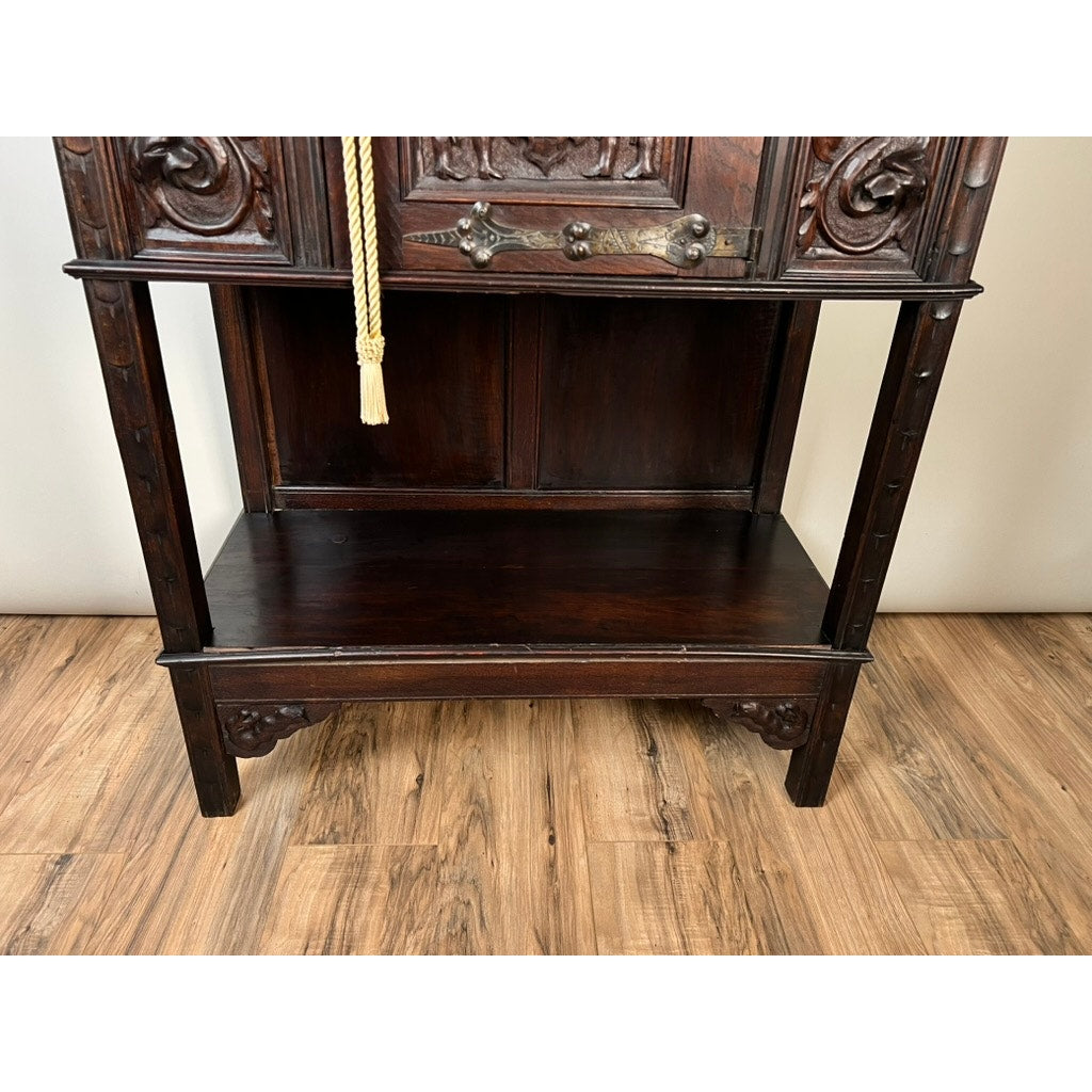 A close-up of the lower half of an Antique 19th-Century French Breton-Style Carved Oak Cabinet featuring deeply carved knights, a gold tassel, and an empty shelf, placed on a wooden floor against a light wall.