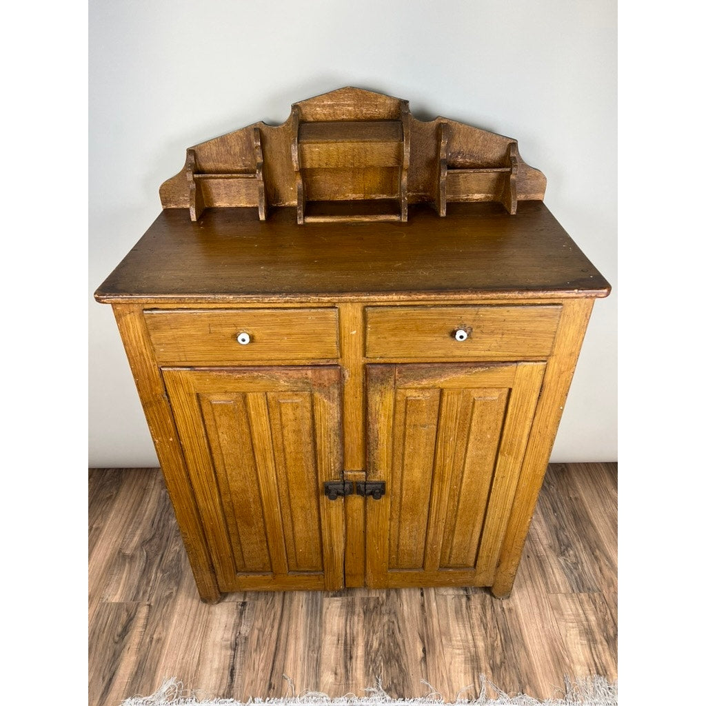The Antique Victorian Country Grain Painted Jelly Cupboard c. 1870 features two paneled doors, two small drawers, and a top tiered shelf, capturing classic vintage charm against a light gray wall and wooden floor. Brand unknown.