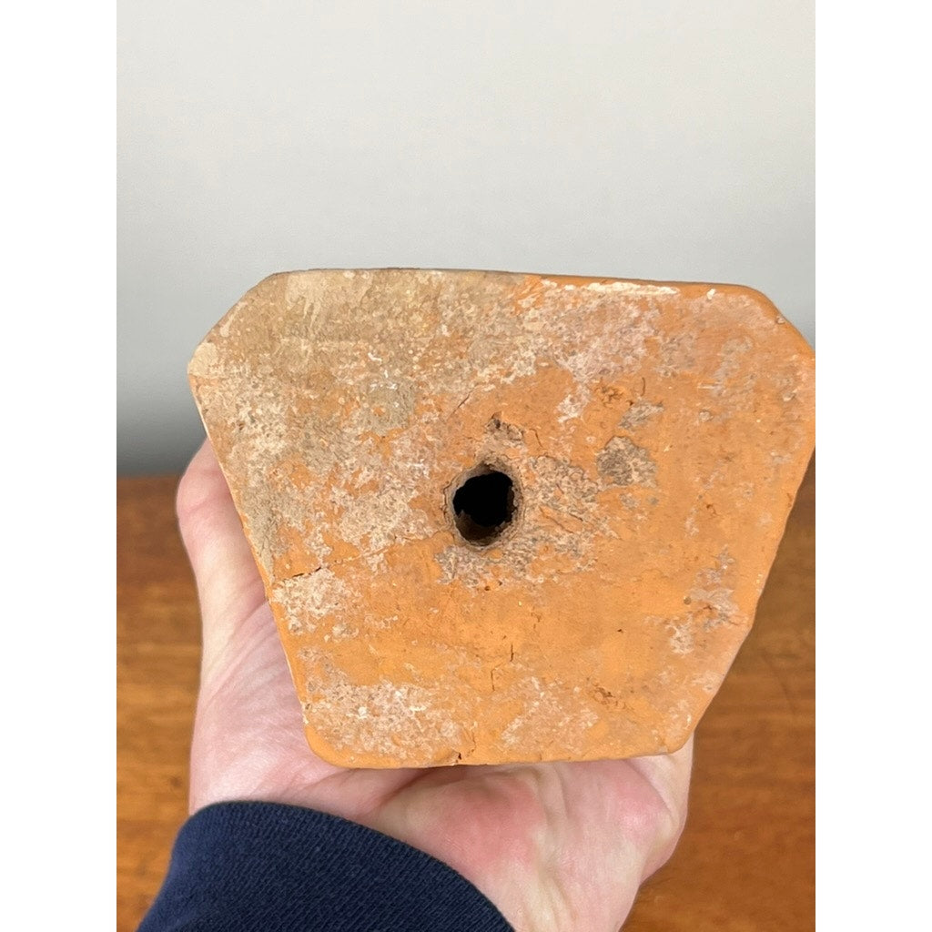 A hand holds the bottom of an Antique Chinese Tang Dynasty (618-907 AD) Terracotta Figure of a Standing Court Lady with COA, showing its textured orange-brown surface and drainage hole against a light background.