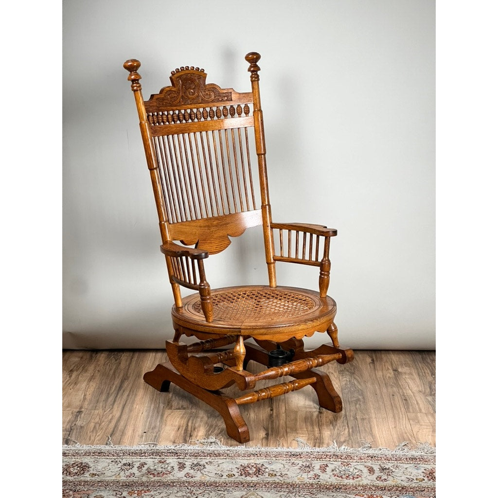 Antique Victorian Oak Platform Rocker (late 19th century) with tall ornate backrest, turned spindles, armrests, and woven cane seat rests on a wooden floor by a patterned rug, set against a plain light background. Brand: Unknown.
