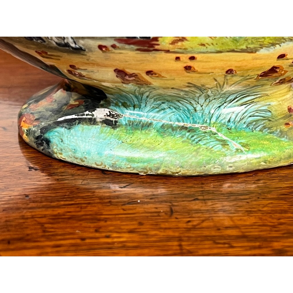 Close-up of the painted base of an Antique Italian Renaissance Revival Hand Painted Maiolica Vase with Winged Putti Handles (c. 1920-1940), featuring green grass, brown spots, and textured detailing, on a wooden surface.